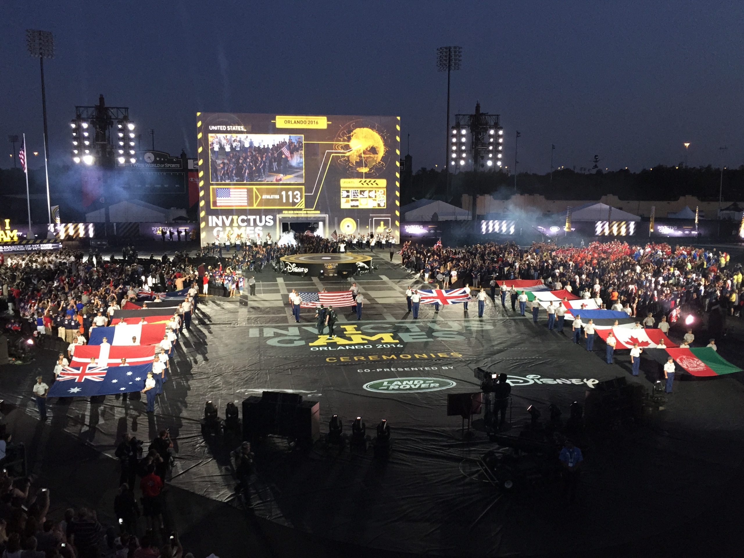 outdoor multicultural sporting event taking place at night with large crowds and a large projection screen and lights in the back. In front is a big black tarp saying that says "Invictus Games ceremonies" and different national flags line the perimeter. US flag and English flag are in the middle of the black tarp.