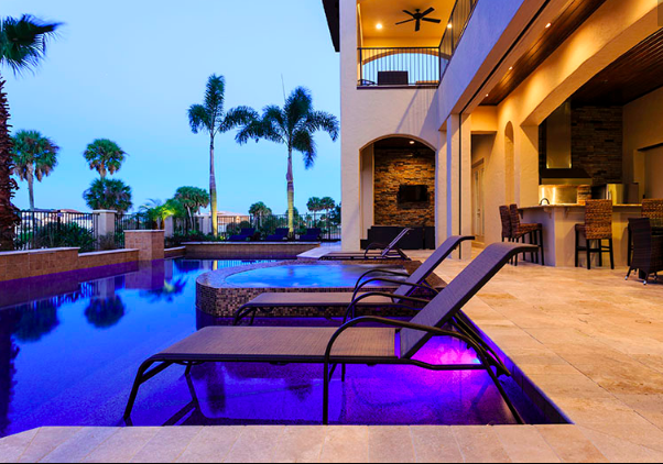 house poolside seating with chaises and purple lighting in the pool and a hot tub in the pool. Palm trees surround the perimeter and the area is gated. White house with lots of open outdoor space and an outdoor kitchen to the right of the pool with island and tall seating.