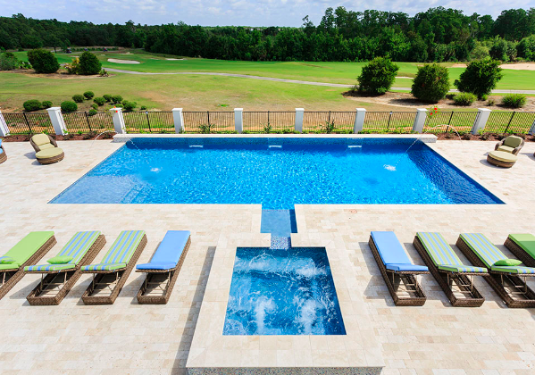 rectangular pool with small hot tub perpendicular and blue and green chaises next to the hot tub, four on each side. Gated area with view of green grass and trees. One chair on either side of the pool facing the chaises