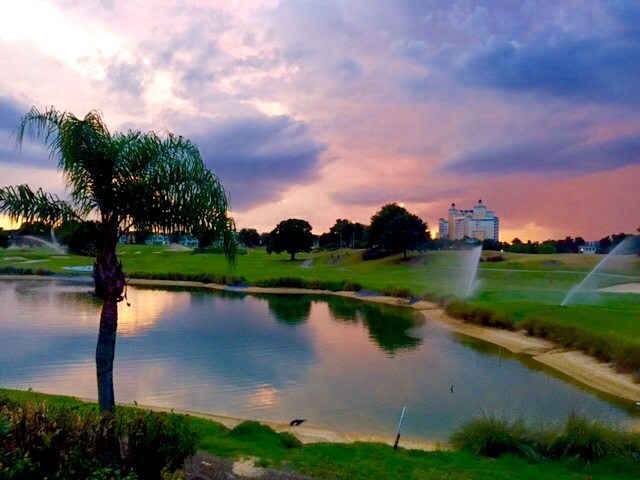 Beautiful purple sunset with clouds on the golf course. Large lake in front and sprinklers on the green. Trees around the area and large building in the background.