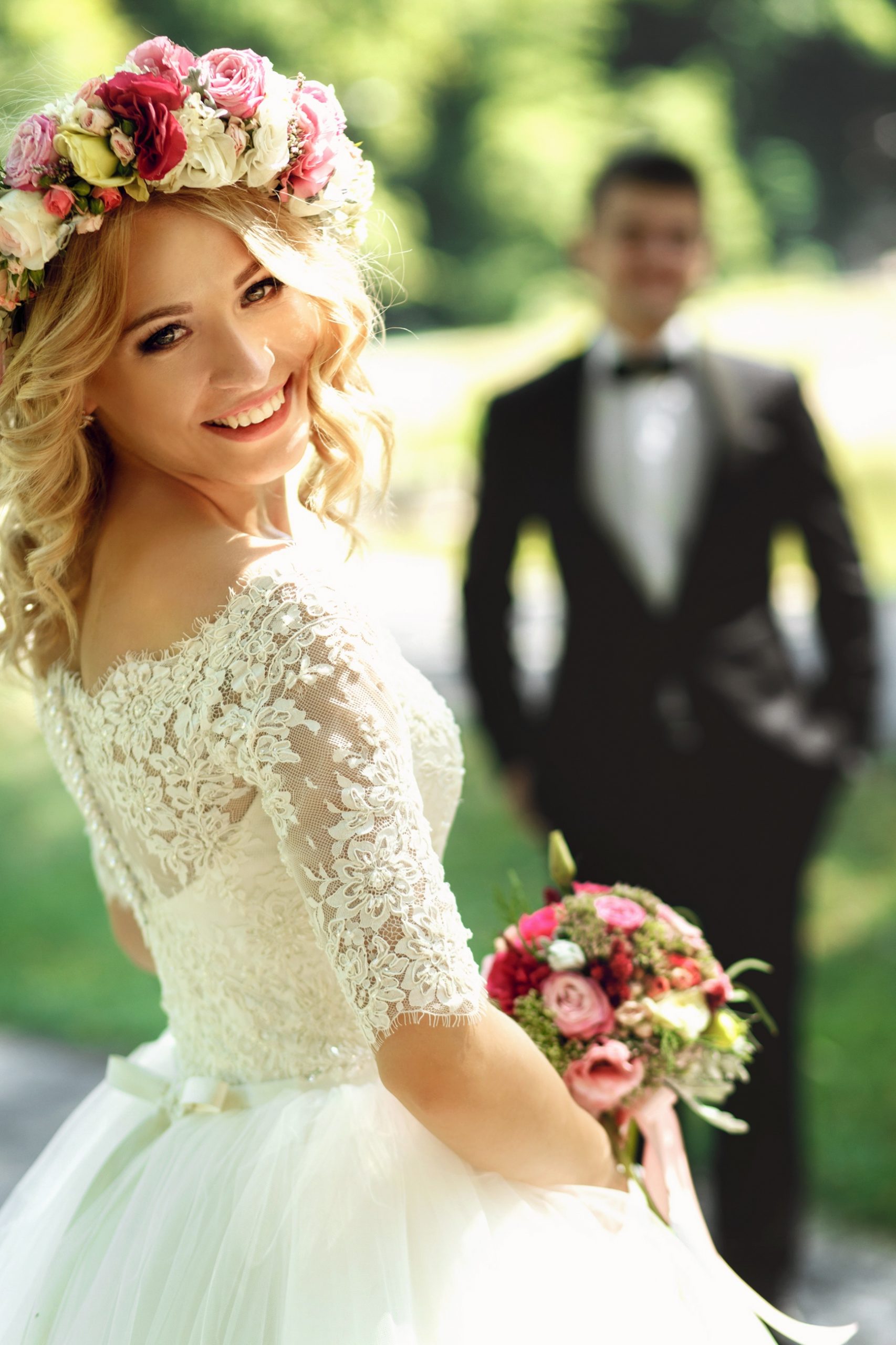 A beautiful bride smiling with her back to the camera. She is wearing a flower crown and holding a matching bouquet and in the background stands the groom.