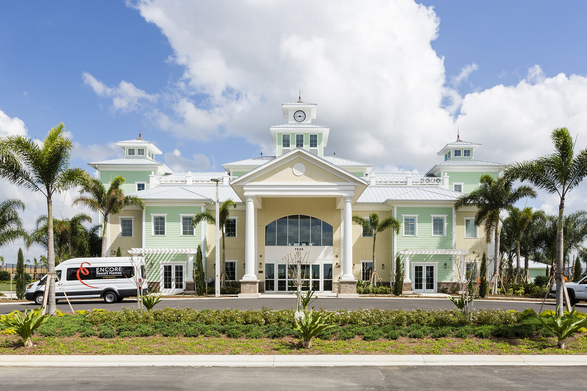 Large pale yellow and green building with large driveway in front and palm trees surrounding. There is a small white bus outside for Encore Resort.
