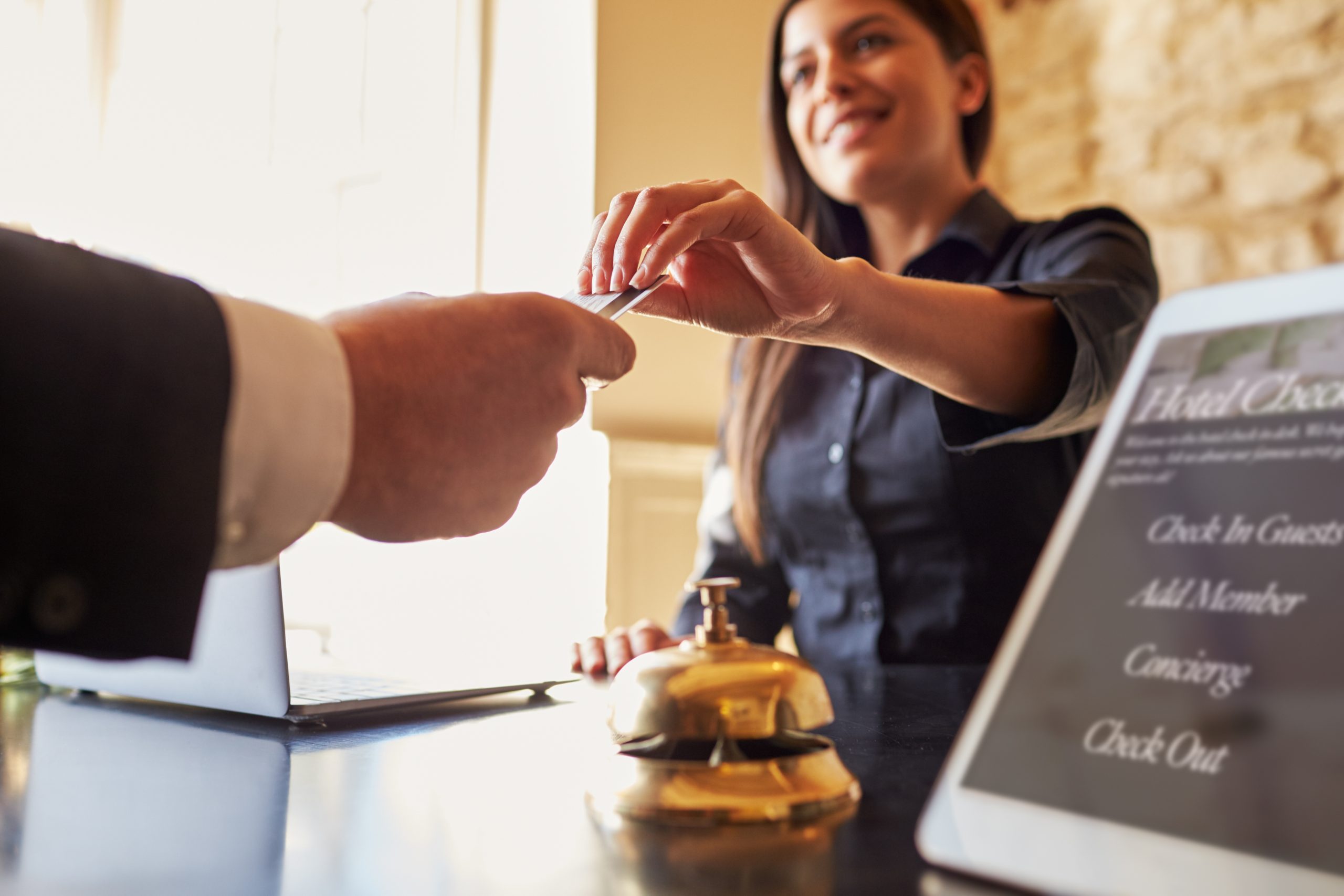 A concierge desk worker is handing a key card to a customer. There is a bell in the photo and a laptop and a sign telling guests to check in and out