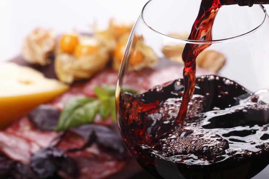 A close up of red wine being poured into a glass. There is a charcuterie board in the background with salami, cheese, and basil.