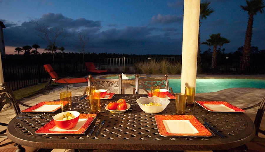 A table set up for dinner by a pool at a house. There are six settings of red plates, glasses, and a bowl of apples and a bowl of salad on the table.
