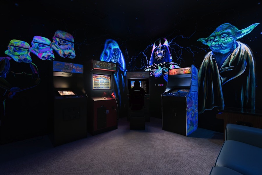 A dark arcade room with four games and glow in the dark images from Star Wars, including Darth Vader and Yoda. One person is playing a game in the corner of the room.