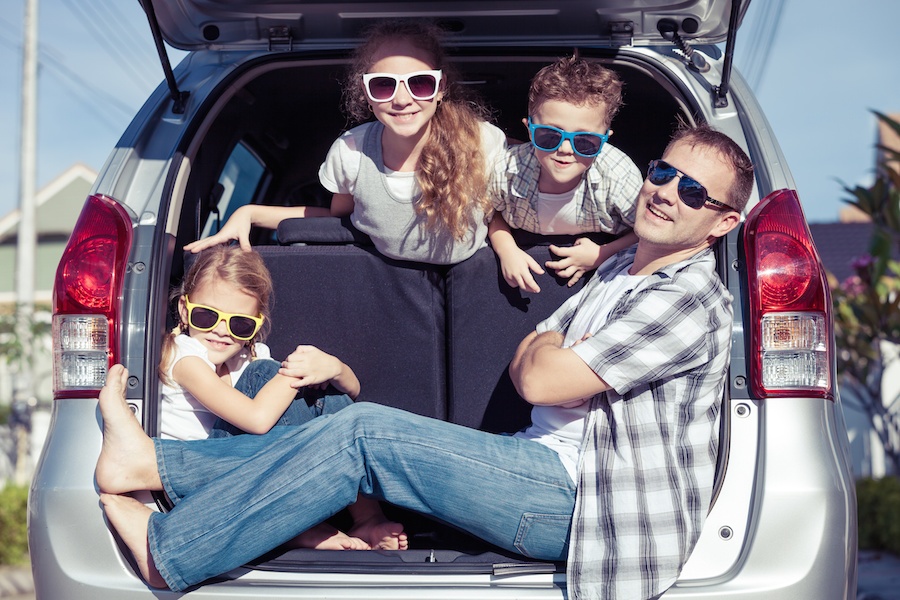 A family is looking out the trunk of their car. They are all wearing sunglasses. A man is sitting with his body spread across the trunk and a young girl is behind him. A young girl and boy are looking over the seat above them.