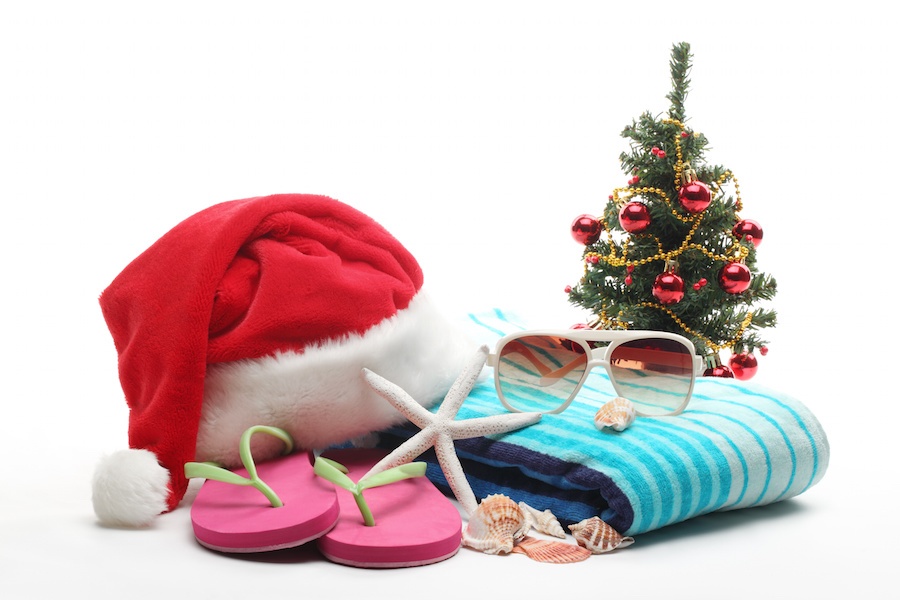 A white background with a blue striped towel, pink and green flip flops, a starfish and shells, sunglasses and a santa hat on top of the towel and a small Christmas tree with large red and yellow ornaments in the back