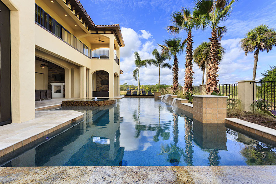 An off white house is to the right of a large rectangular pool with palm trees lining the perimeter and a gate. The pool has brick along the righthand side and waterfalls coming from the brick.