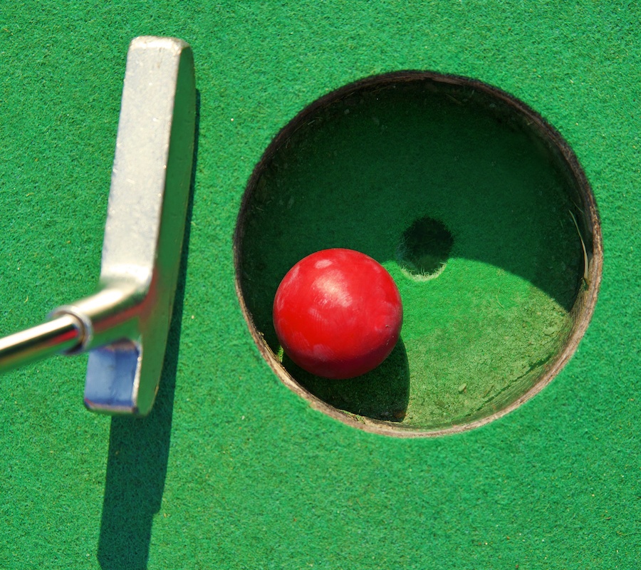 A close up of a putter hitting a red golf ball into a hole