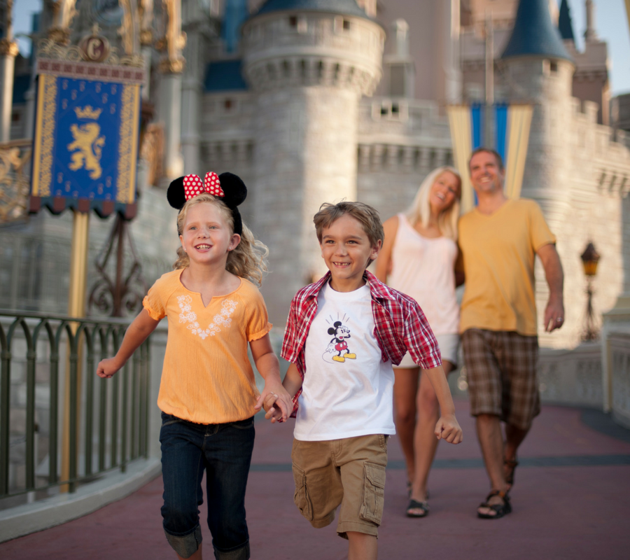 A young boy and young girl wearing mini mouse ears are holding hands and running away from the Disney castle while their parents walk behind them