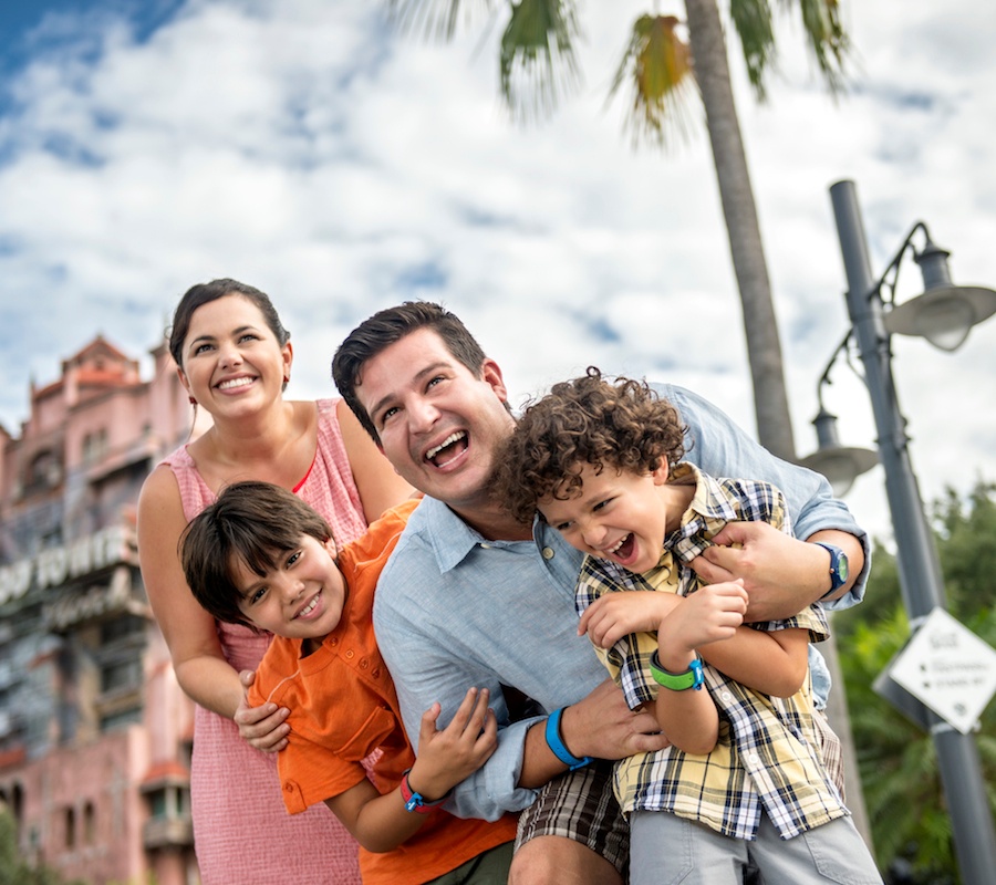 A family of two boys, the father is hugging one of the boys from behind and then the other boy is hugging the dad from behind while the mother stands behind