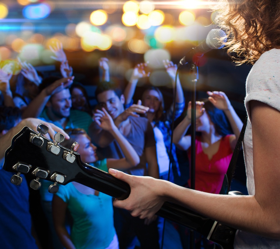 A woman playing guitar and singing into a microphone in front of a small crowd of people with their hands in the air