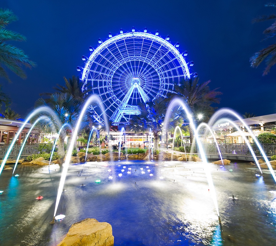 An image of a body of water with sprinklers all around shooting into the middle and palm trees in the background. There is a large blue light up ferris wheel behind.