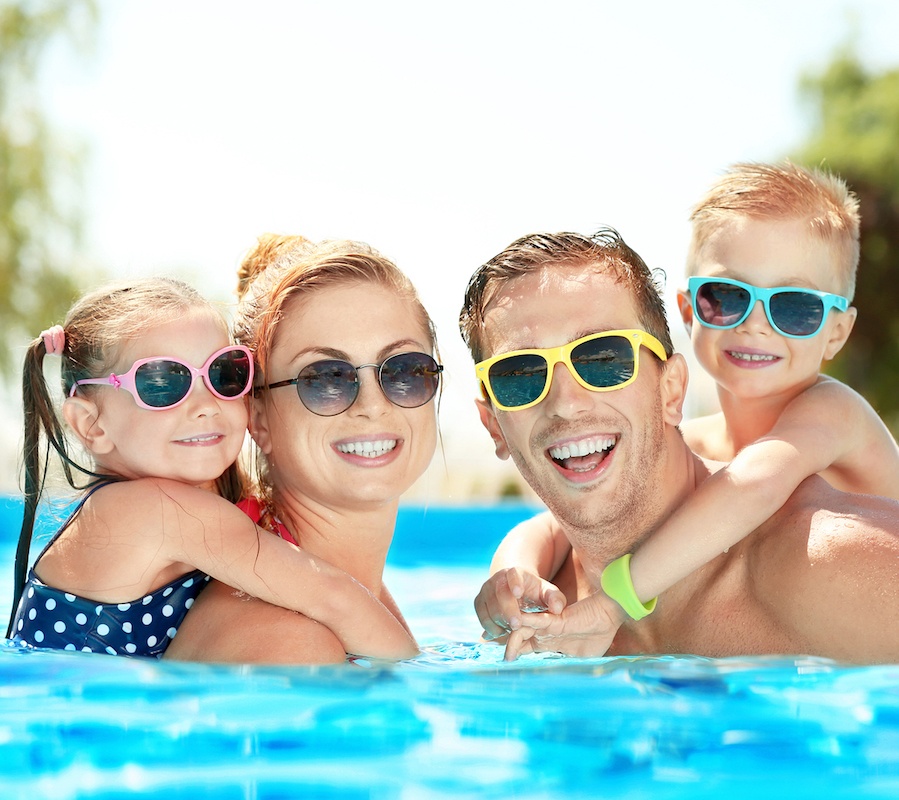 A family wearing sunglasses in the pool. On the left, a little girl is on her mother's back. On the right, a little boy is on his father's back.