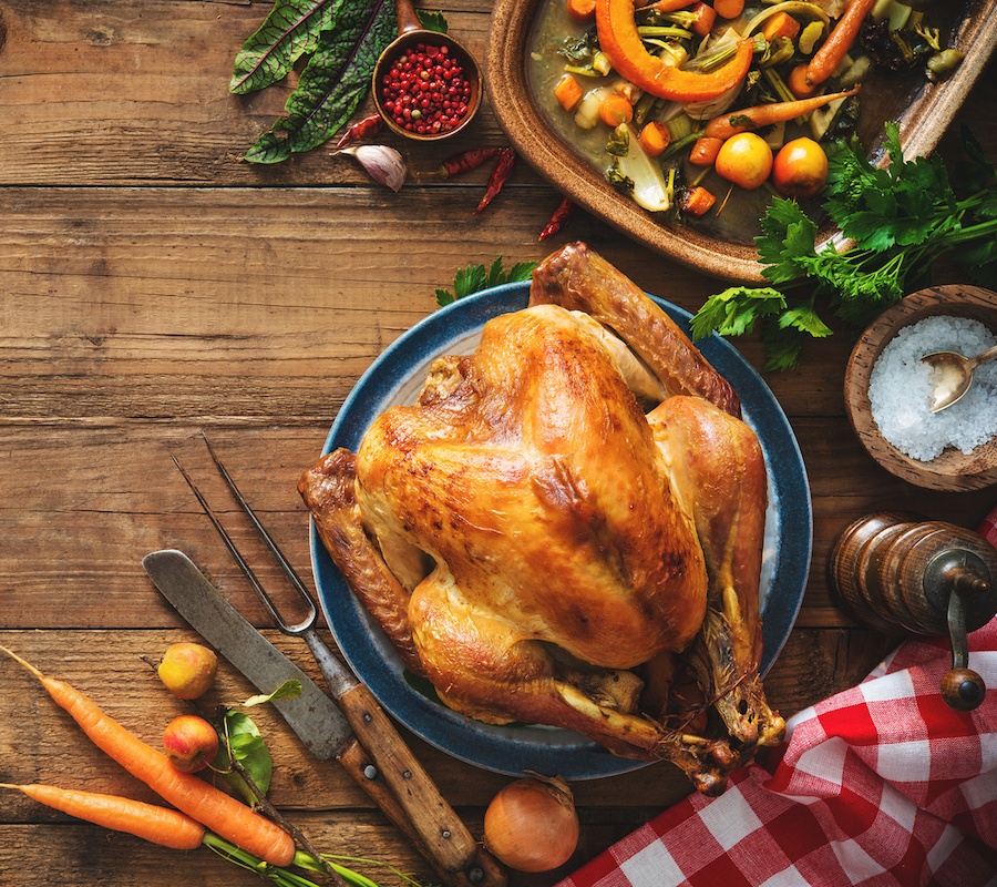 A close up picture of a roast chicken on a wooden table with a plaid table cloth in the corner and a bowl of salt, carrots, parsley, a stew of vegetables above, and a large serving fork and knife