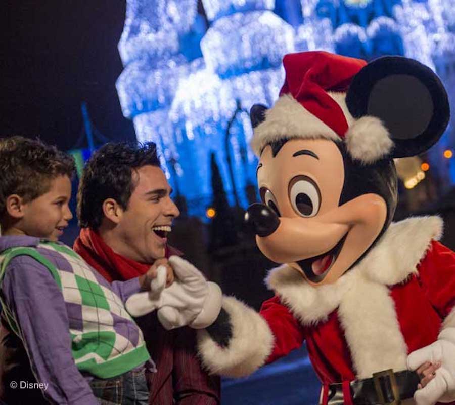 Mickey mouse in a Santa costume is shaking hands with a little boy and his father in front of a snow covered castle