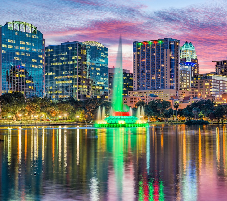 A colorful skyline picture over the water with a large green fountain in the middle squirting water and trees surrounding the area