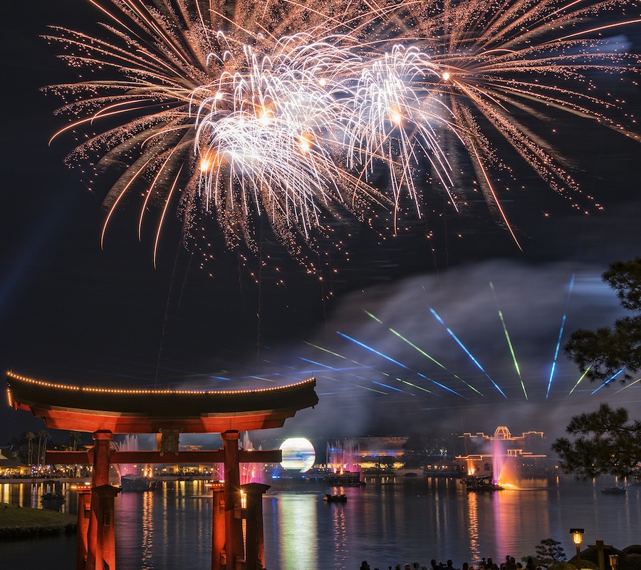 Fireworks over the water. In the front there is a red awning and in the back you can see blue and green streaks of light and boats are giving out light as well