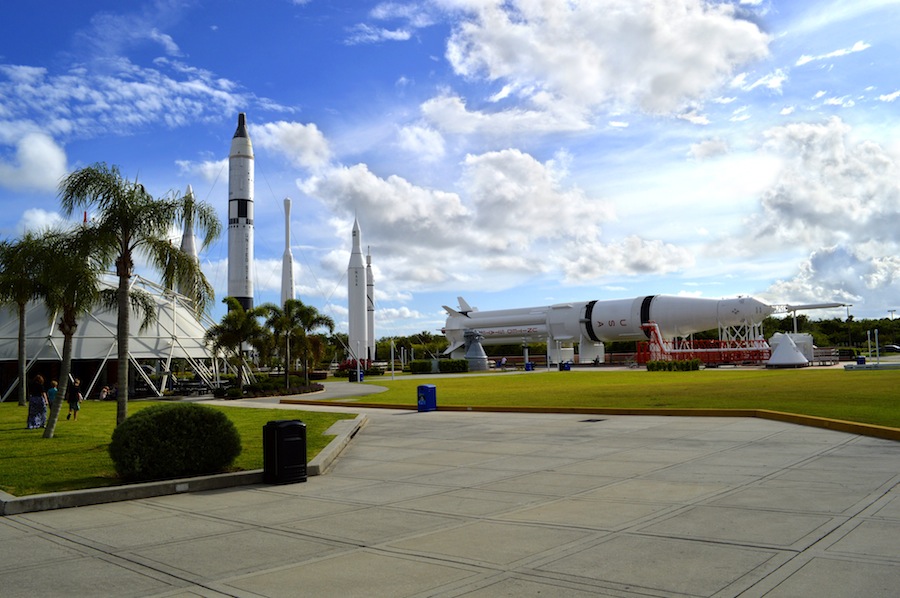 A model space shuttle is set up along with other life size model rockets with green grass surrounding and a sidewalk leading over to the space area
