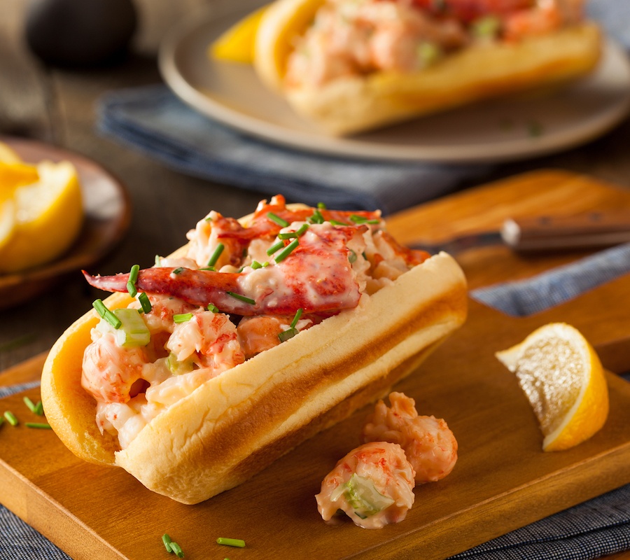 A close up picture of a lobster roll on a wooden cutting board with a sliced lemon on the side