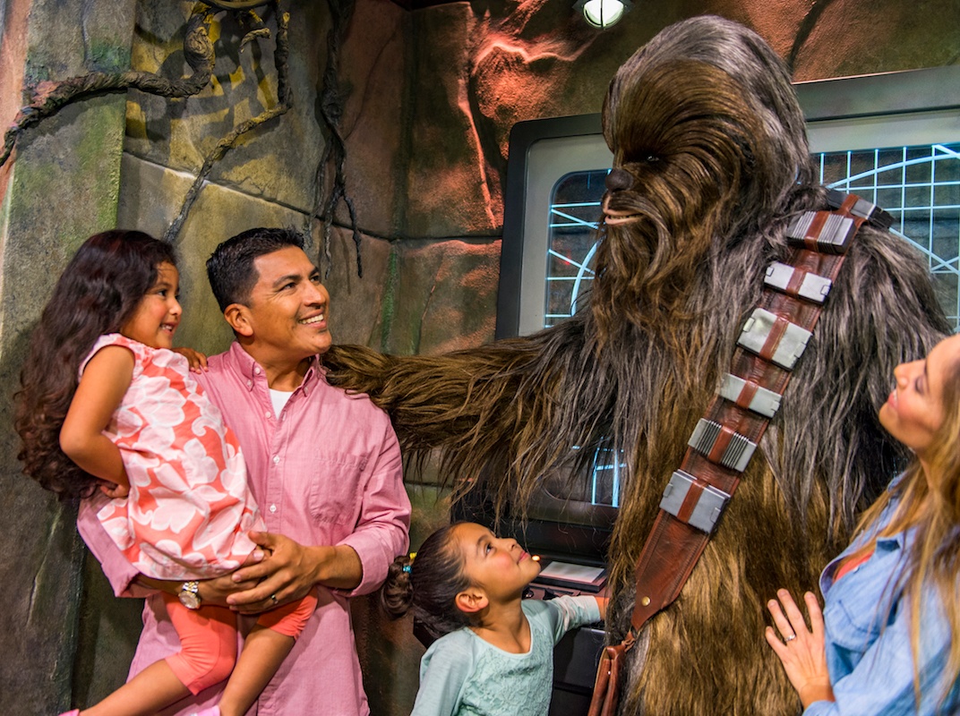 A family meeting Chewbacca: the mom is standing to his right, and two his left is one little girl standing looking up and the father holding up his other daughter