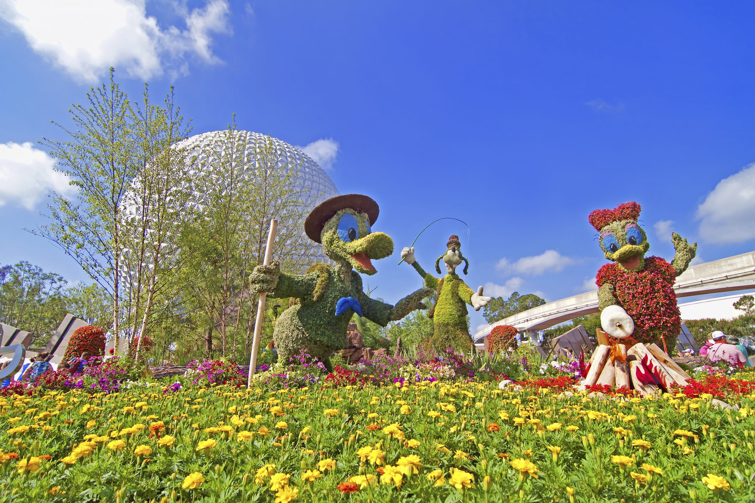 A garden of yellow flowers and tall bushes shaped like Donald and Daffy Duck and Goofy with the Epcot ball behind them to the left