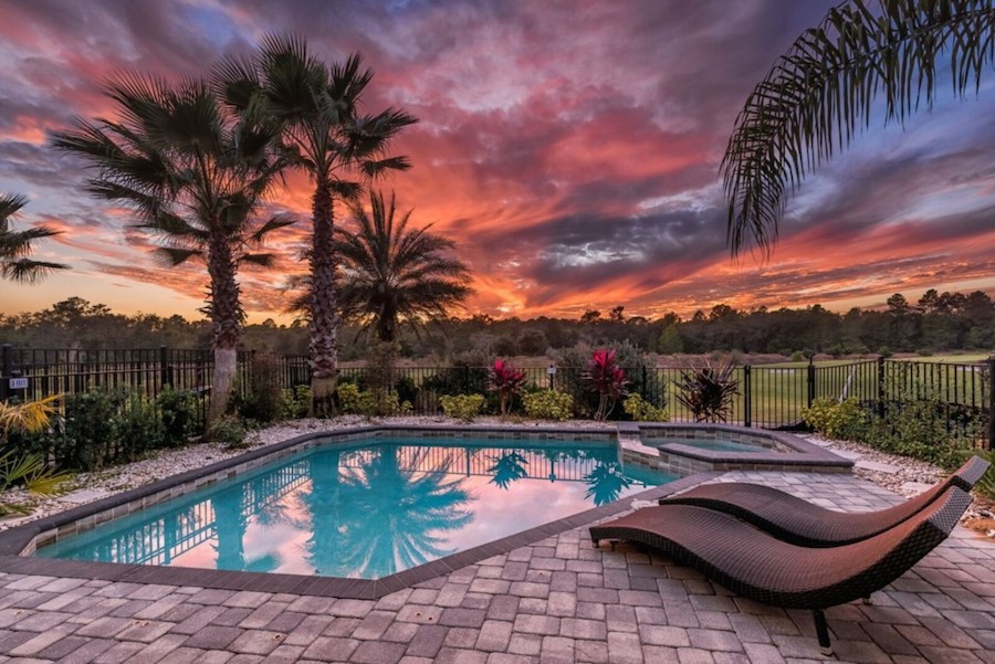 A small pool with attached hot tub with two brown chaises and brick patio. There is a black gate around the pool and palm trees and bushes and pink flowers. There is a beautiful sunset in the back.