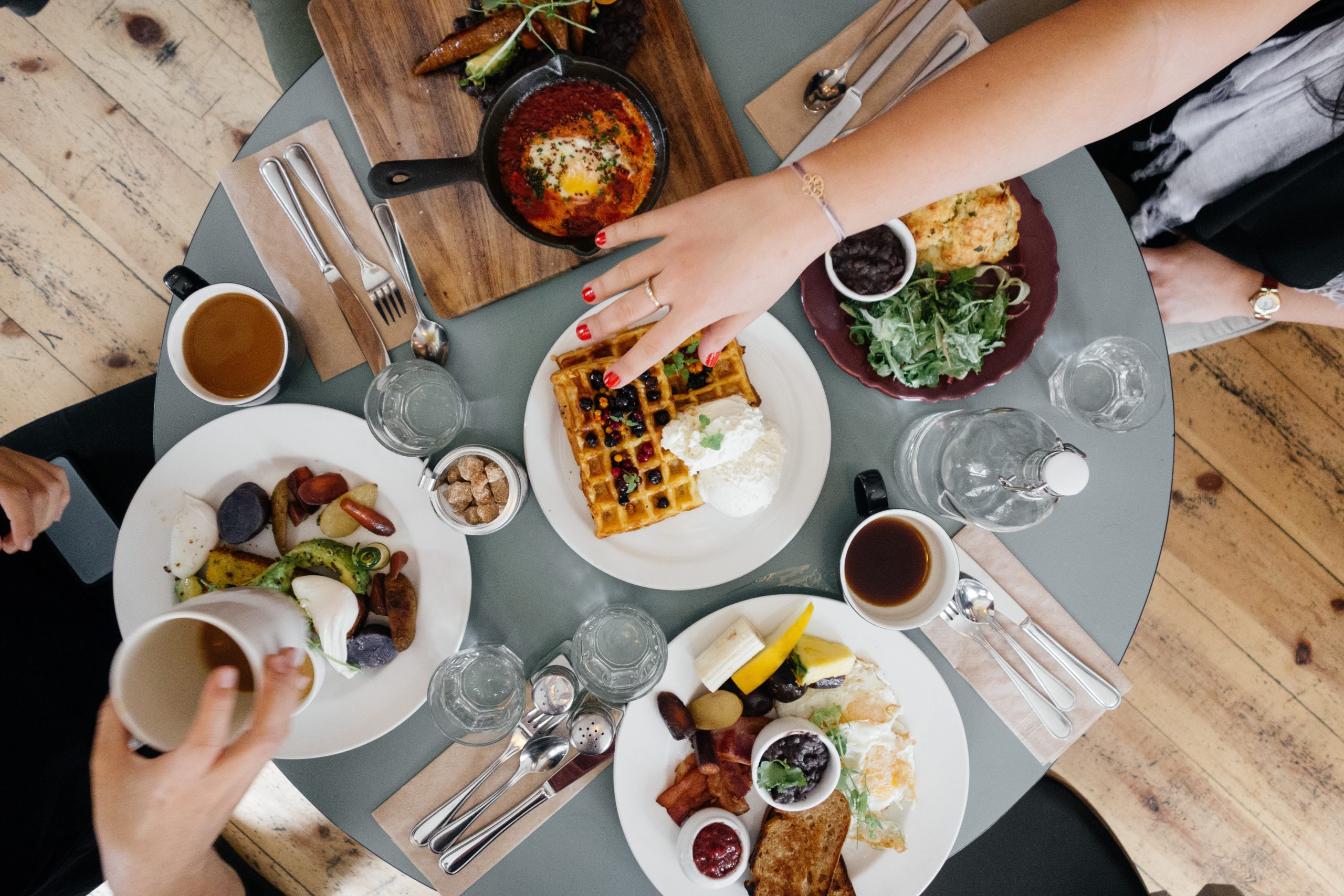 A picture of people's hands reaching over a breakfast table featuring coffee, water, waffles with fruit and whipped cream, greens, toast, eggs, bacon, ketchup, shakshuka, silverware, and more