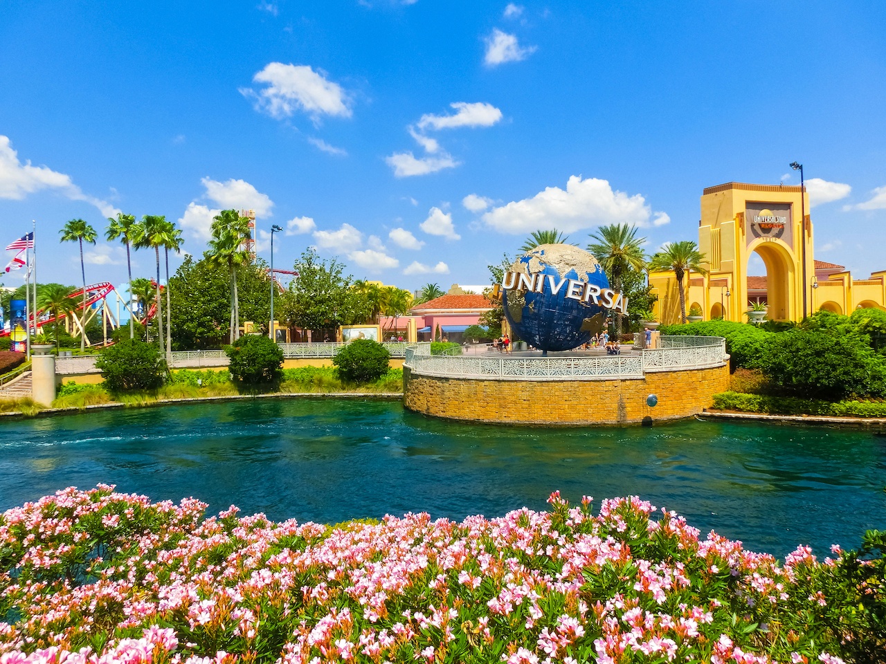 A picture of the universal globe surrounded by water and pink flowers and bushes and palm trees. In the back you can see the entrance to the park