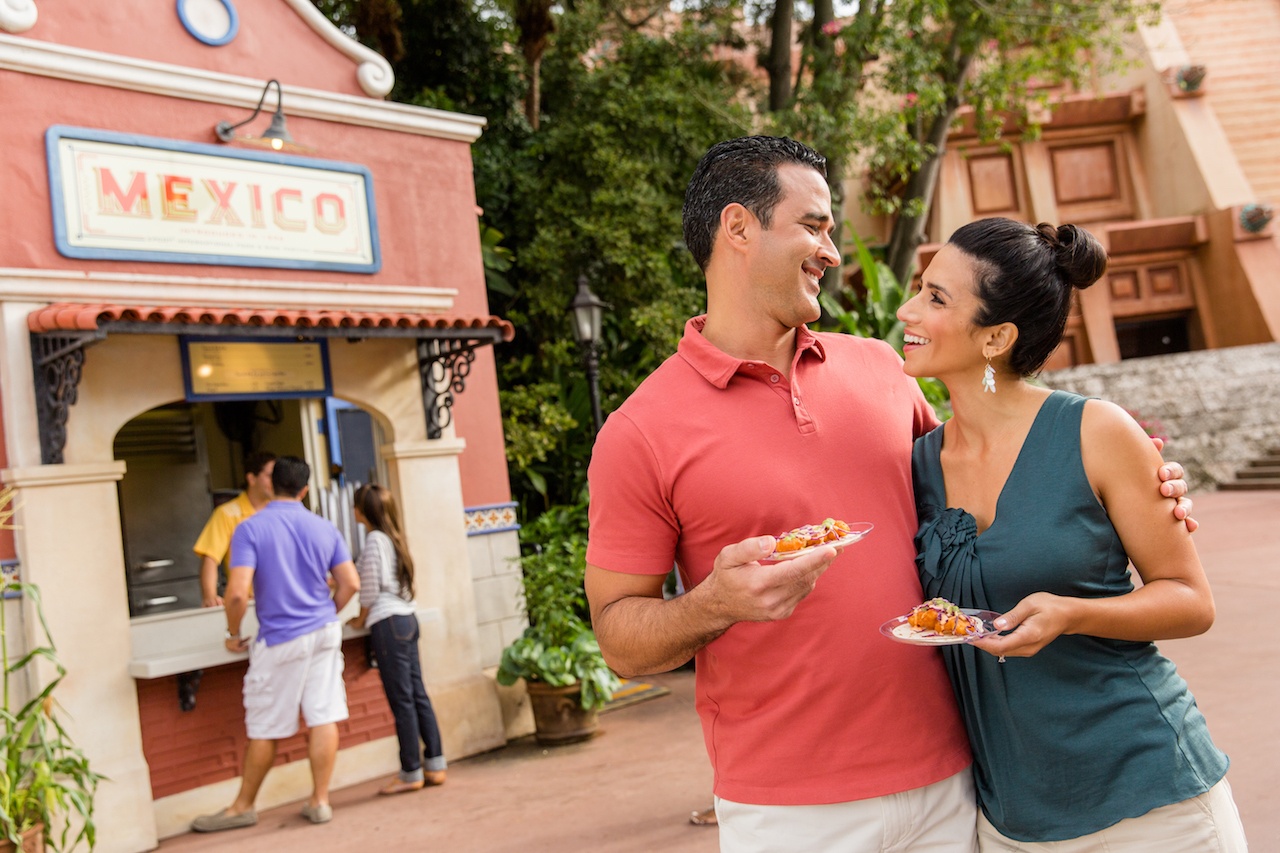 A man and a woman are holding each other and smiling in front of a red food stand that says "Mexico" and holding small plates of food