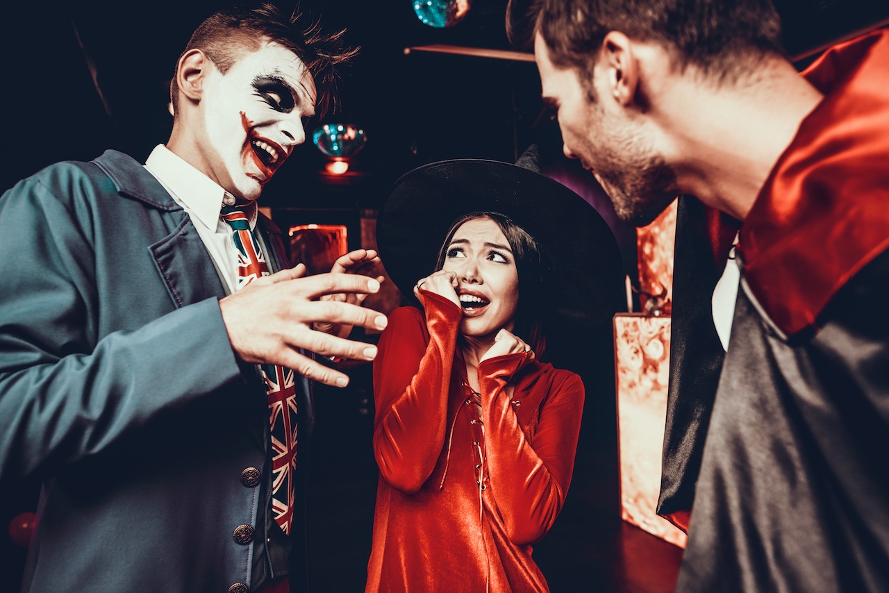 A halloween photo of a man with Joker face paint to the left scaring a girl in a witch hat. A man in a vampire cape is looking at her to the right.