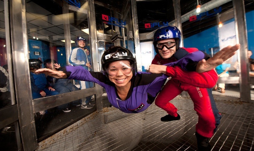 A woman with a helmet and goggles at iFly, where she is in the air with her arms out and a man who is also wearing a helmet and goggles is holding her