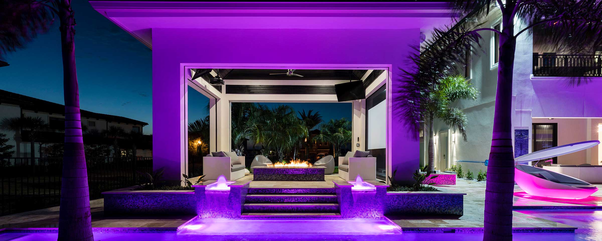 Purple pool lights on in the evening surrounding cabana with wood burning fire in the center.